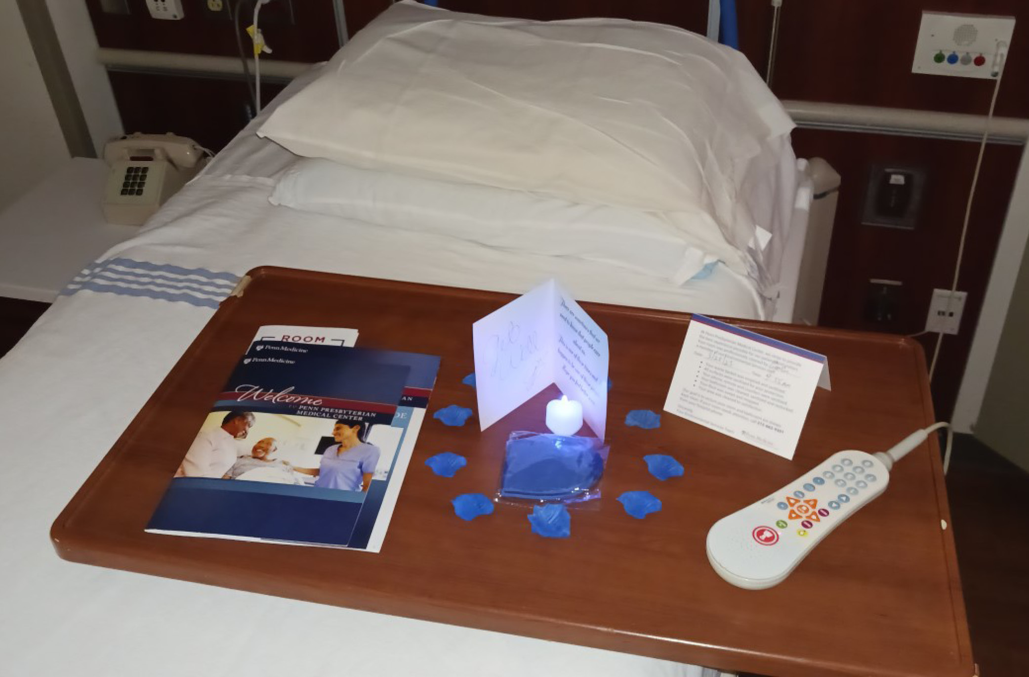 Objects on a hospital bedside table include a get-well card, blue fabric flower petals, and a glowing, battery-powered candle 